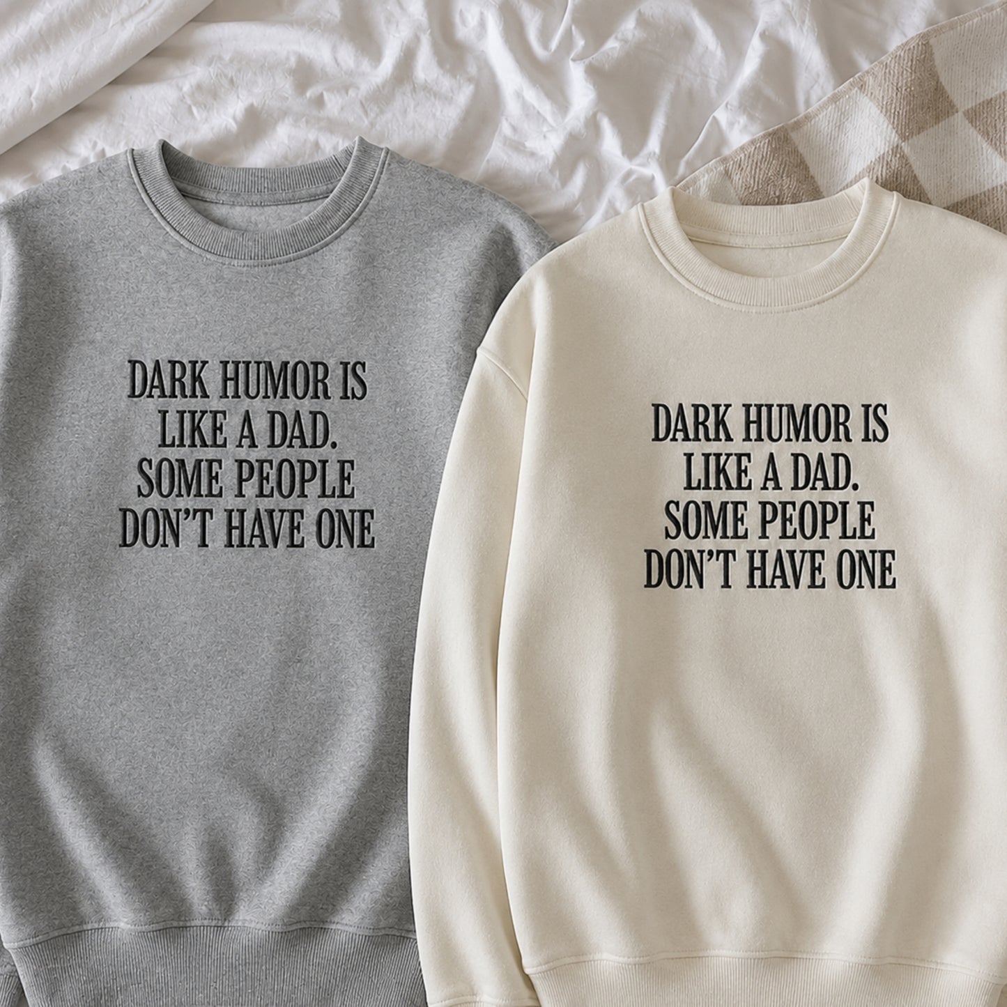 Two crewneck sweatshirts laid flat on white bedding. One is heather gray with black text reading 'DARK HUMOR IS LIKE A DAD.