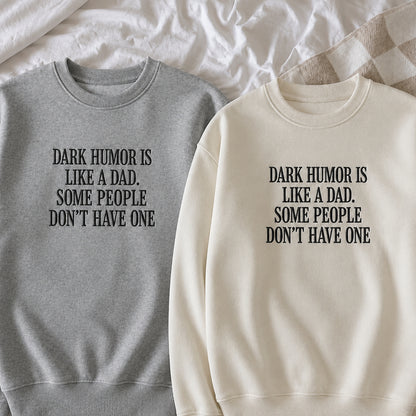 Two crewneck sweatshirts laid flat on white bedding. One is heather gray with black text reading 'DARK HUMOR IS LIKE A DAD.
