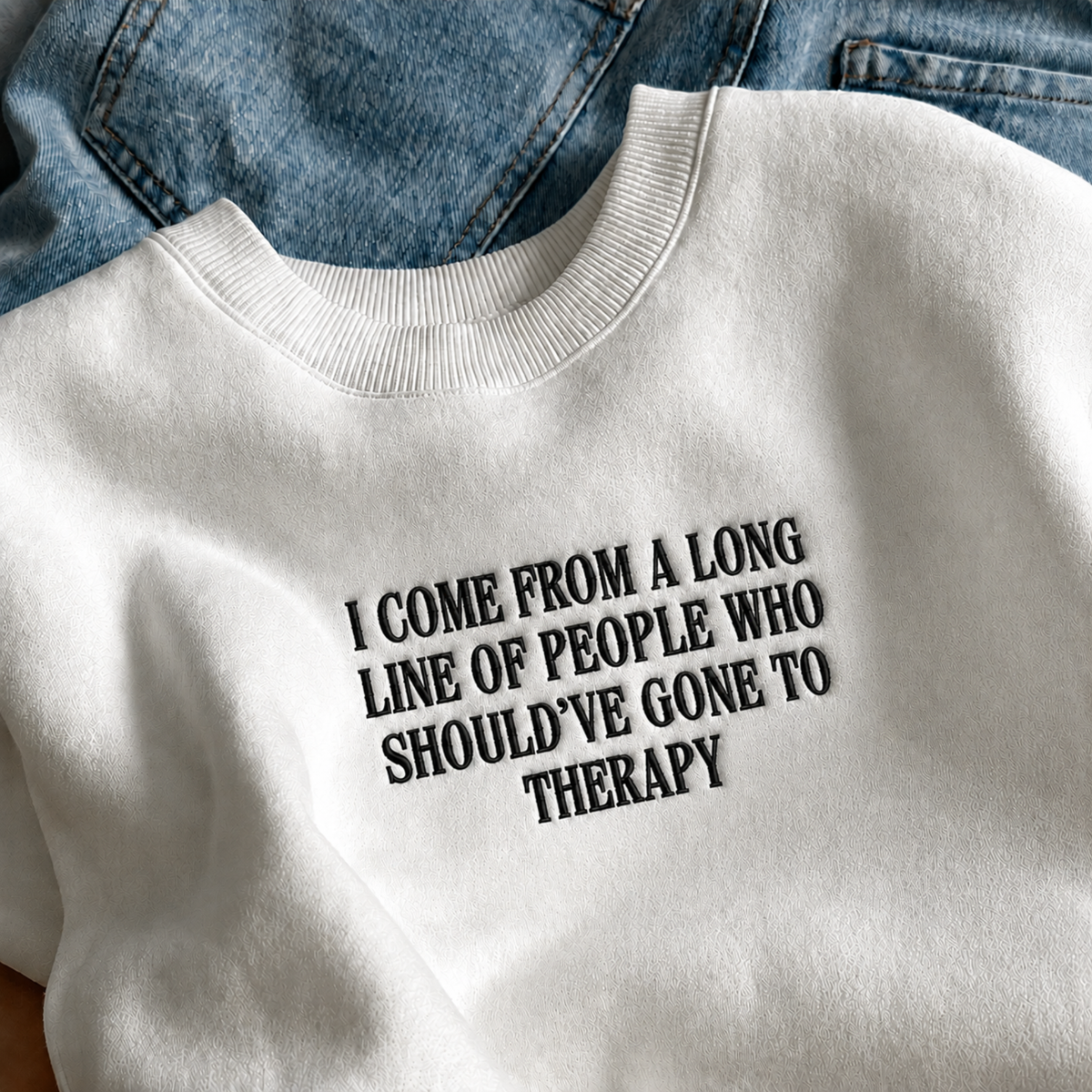 A white crewneck sweatshirt with black text reading 'I COME FROM A LONG LINE OF PEOPLE WHO SHOULD'VE GONE TO THERAPY', styled flat-lay on top of blue denim jeans.