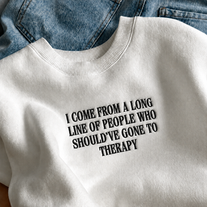 A white crewneck sweatshirt with black text reading 'I COME FROM A LONG LINE OF PEOPLE WHO SHOULD'VE GONE TO THERAPY', styled flat-lay on top of blue denim jeans.