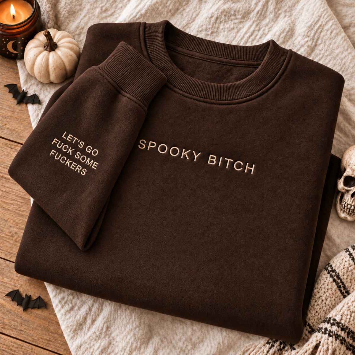 A folded dark brown crewneck sweatshirt with gold embroidered text reading 'SPOOKY BITCH' on the chest and 'LET'S GO FUCK SOME FUCKERS' on the sleeve, styled on a textured cream blanket with Halloween props including a candle, small pumpkin, bats, and a skull.