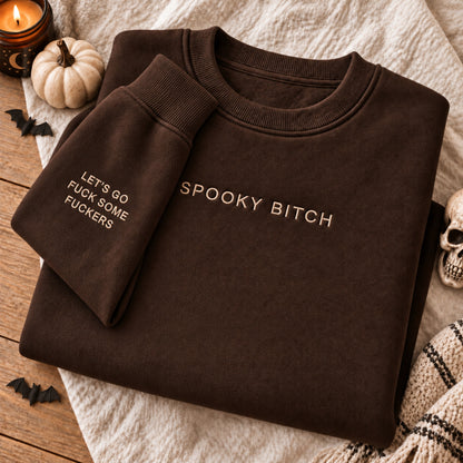 A folded dark brown crewneck sweatshirt with gold embroidered text reading 'SPOOKY BITCH' on the chest and 'LET'S GO FUCK SOME FUCKERS' on the sleeve, styled on a textured cream blanket with Halloween props including a candle, small pumpkin, bats, and a skull.