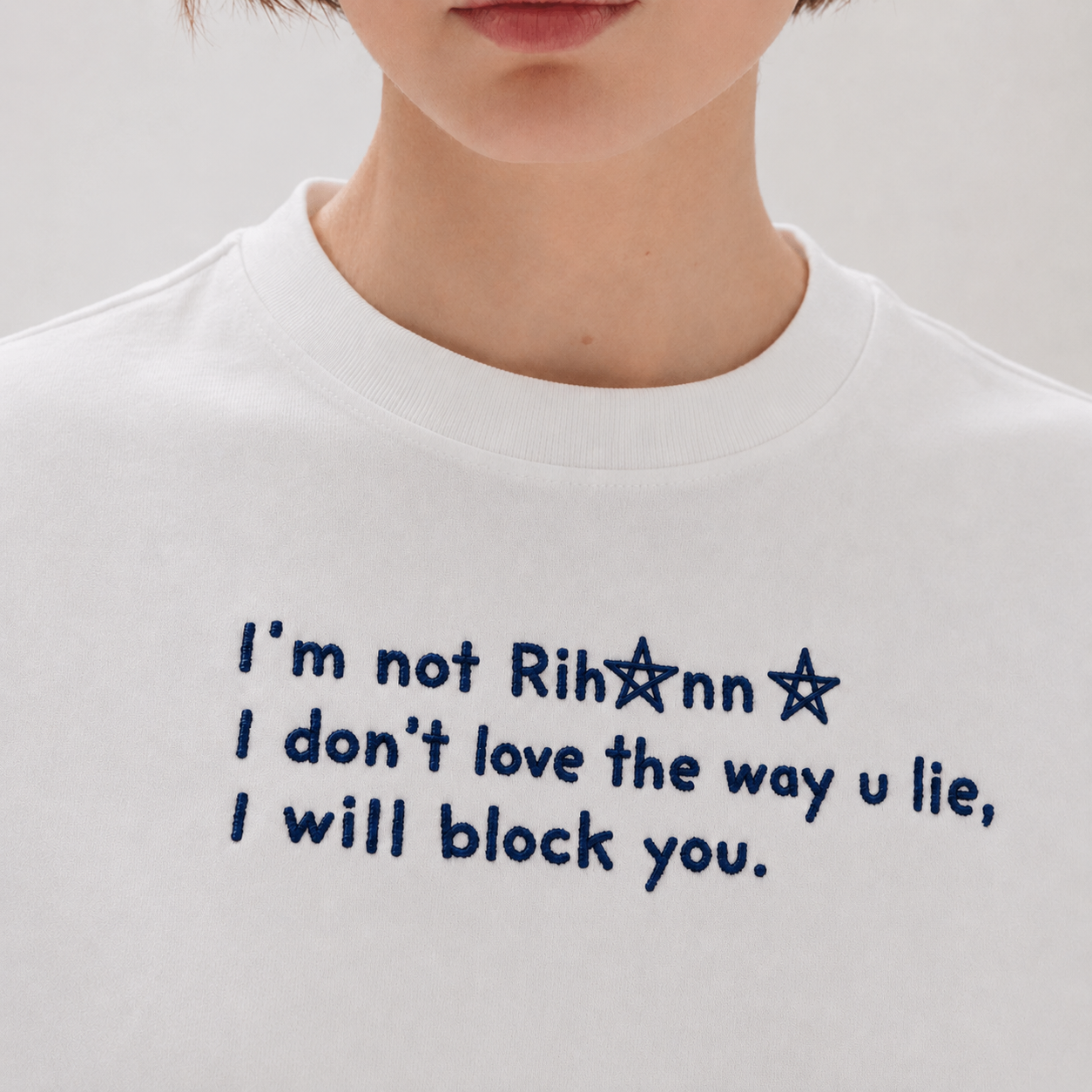 Close-up of a person wearing a white shirt embroidered with blue text reading "I'm not Rihanna I don't love the way u lie, I will block you.