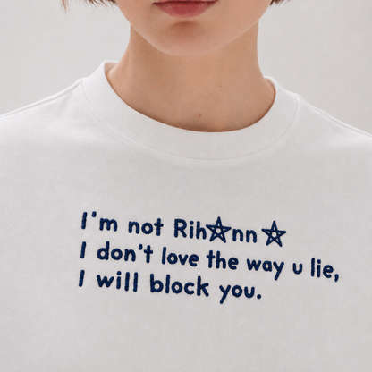 Close-up of a person wearing a white shirt embroidered with blue text reading "I'm not Rihanna I don't love the way u lie, I will block you.