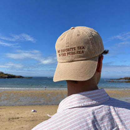 A man viewed from behind wearing a beige baseball cap with the embroidered text 'MY FAVORITE SEA IS THE PUSS-SEA' on a sunny beach with blue sky and ocean in the background.