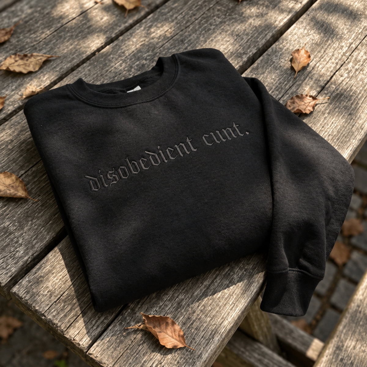 A black crewneck sweatshirt with the phrase 'disobedient cunt.' printed in a gothic-style font across the chest, laid flat on a weathered wooden table with scattered autumn leaves.