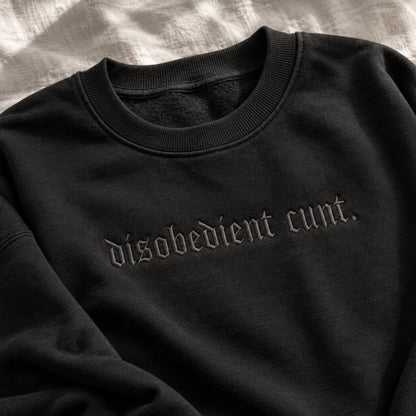 A close-up of a black crewneck sweatshirt featuring the phrase 'disobedient cunt.' embroidered in a gothic font across the chest, laid flat on a textured white fabric background.