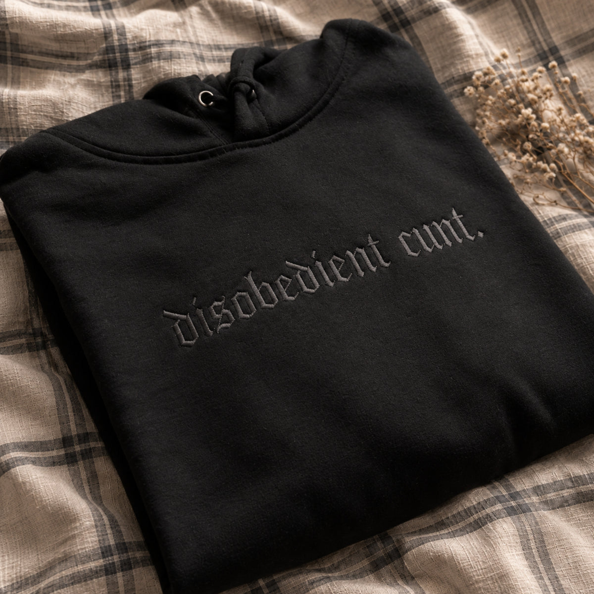 A folded black hoodie with gothic-style embroidered text reading 'disobedient cunt.' lies on a beige plaid fabric surface with dried flowers in the corner.