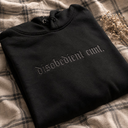 A folded black hoodie with gothic-style embroidered text reading 'disobedient cunt.' lies on a beige plaid fabric surface with dried flowers in the corner.