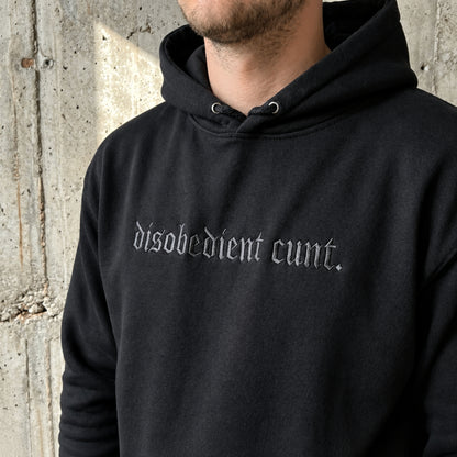 A close-up of a person wearing a black pullover hoodie with the phrase 'disobedient cunt.' embroidered across the chest in a gothic-style light gray font, set against a textured concrete wall.