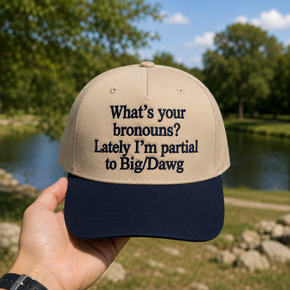 A beige baseball cap with a navy blue brim, held by a hand against a blurred outdoor background of trees and a pond. The front of the cap features the embroidered text: 'What's your bronouns? Lately I'm partial to Big/Dawg'.