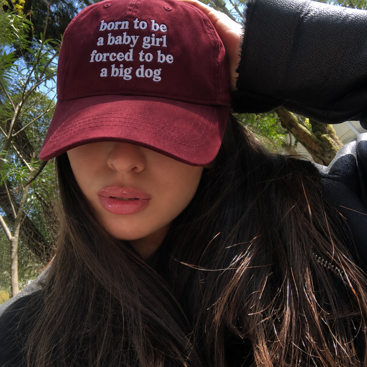 A person outdoors wearing a burgundy baseball cap with white embroidered text that reads 'born to be a baby girl forced to be a big dog'.