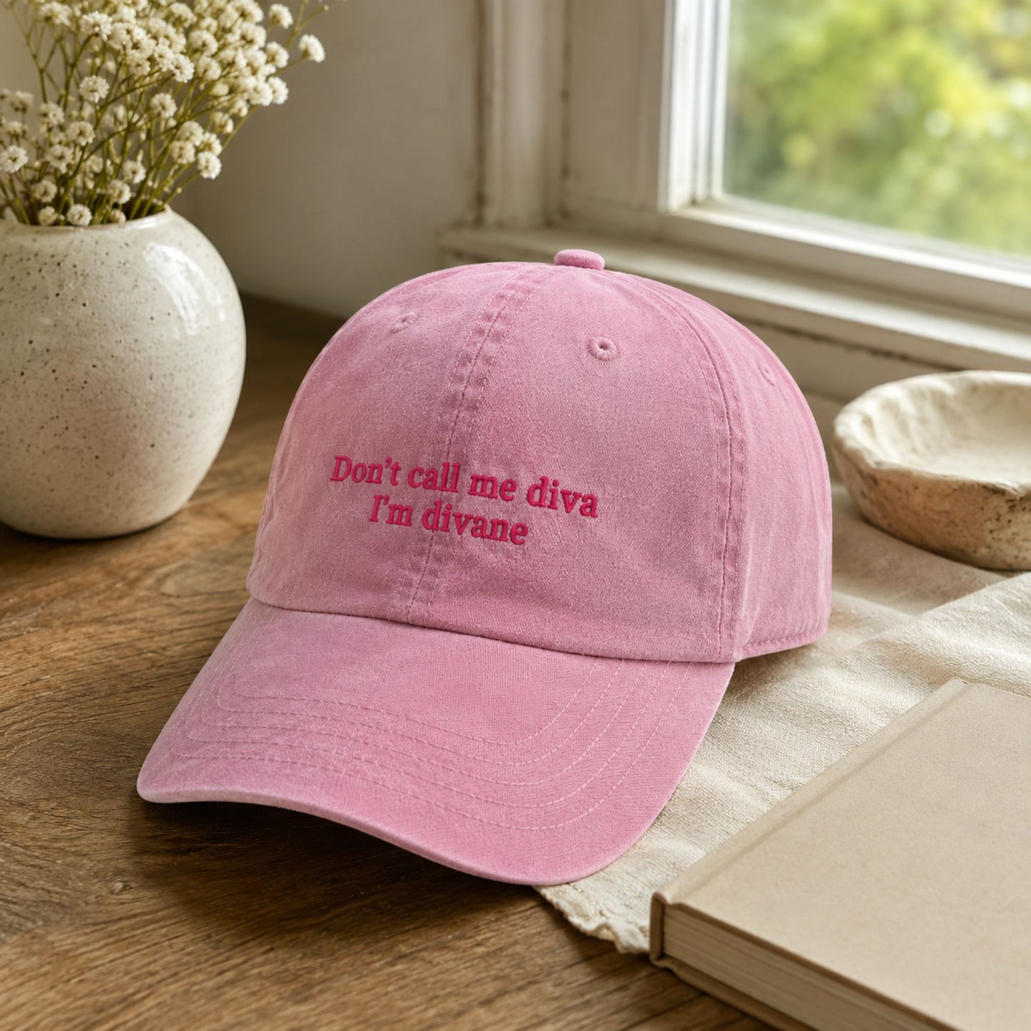 A pink cotton baseball cap with embroidered text reading 'Don't call me diva I'm divane' in a darker pink/red thread, resting on a wooden table with a book, ceramic bowl, and vase of flowers nearby.