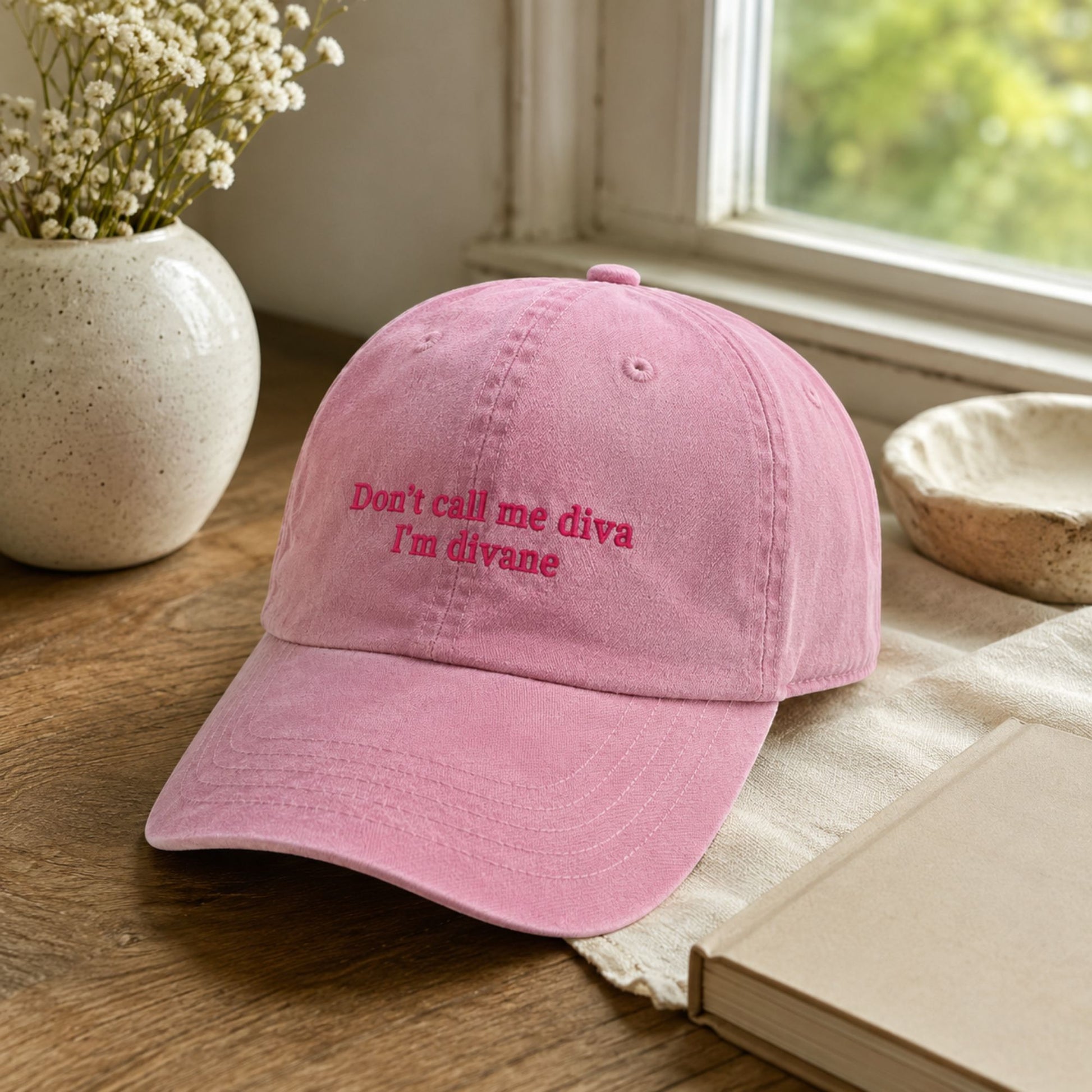 A pink cotton baseball cap with embroidered text reading 'Don't call me diva I'm divane' in a darker pink/red thread, resting on a wooden table with a book, ceramic bowl, and vase of flowers nearby.