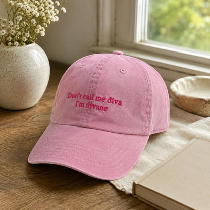 A pink cotton baseball cap with embroidered text reading 'Don't call me diva I'm divane' in a darker pink/red thread, resting on a wooden table with a book, ceramic bowl, and vase of flowers nearby.