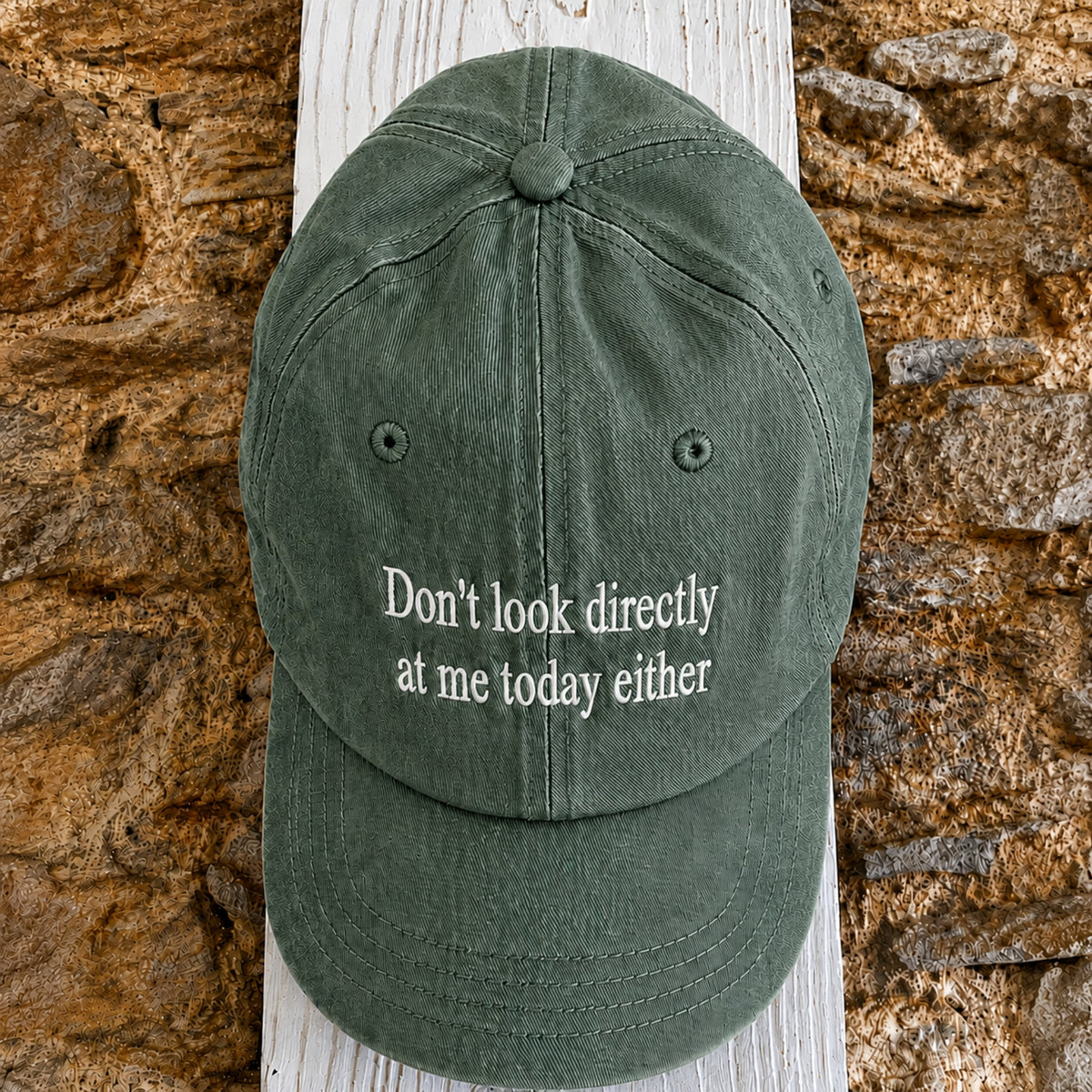 An olive green baseball cap with the phrase 'Don't look directly at me today either' embroidered in white on the front, displayed on a white wooden post against a rustic stone wall.