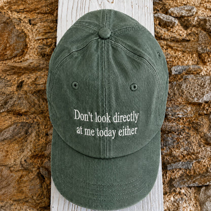 An olive green baseball cap with the phrase 'Don't look directly at me today either' embroidered in white on the front, displayed on a white wooden post against a rustic stone wall.