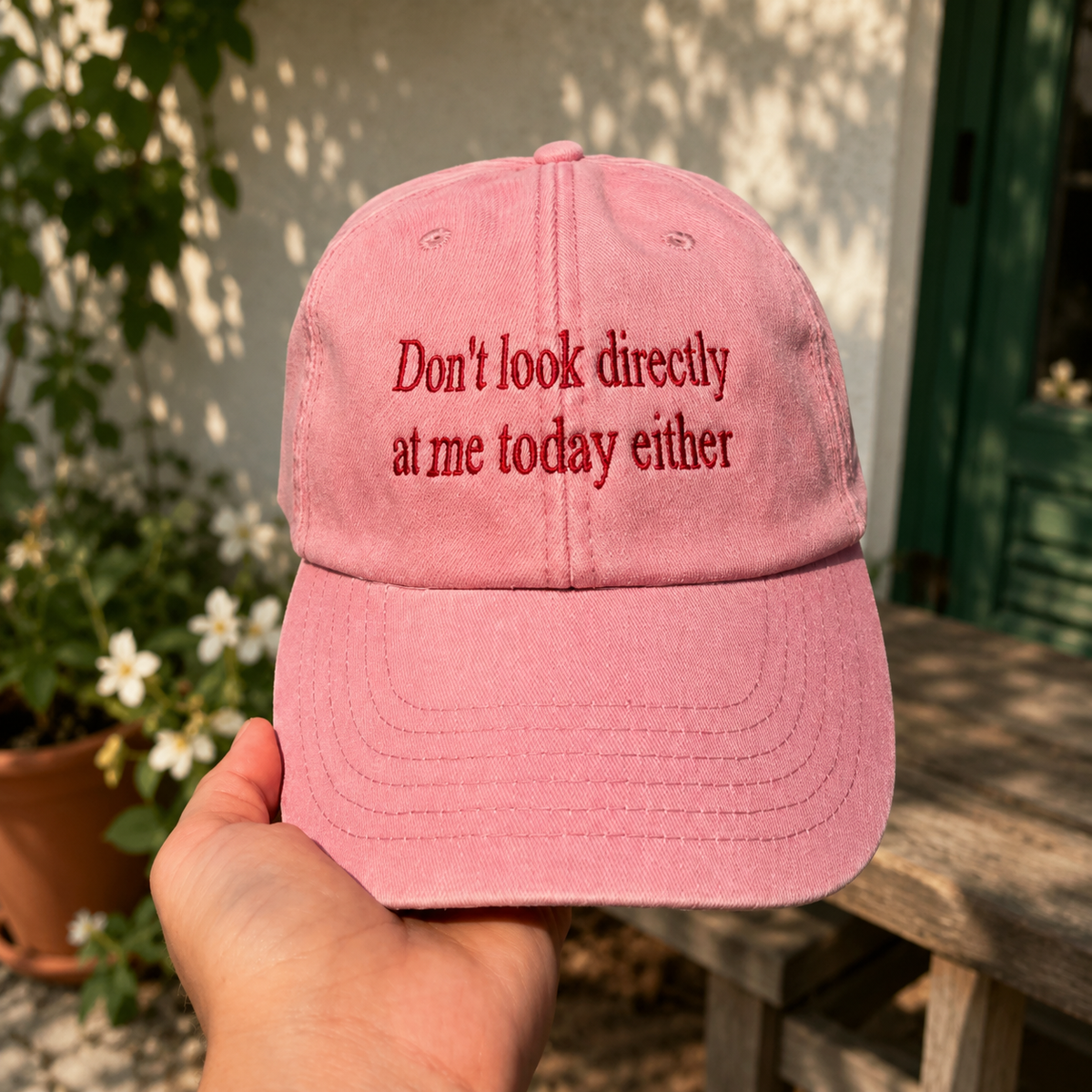 A pink baseball cap held by a hand, featuring the embroidered text 'Don't look directly at me today either' in a darker red font. The background shows a sunny outdoor patio setting with a wooden table and potted plants.