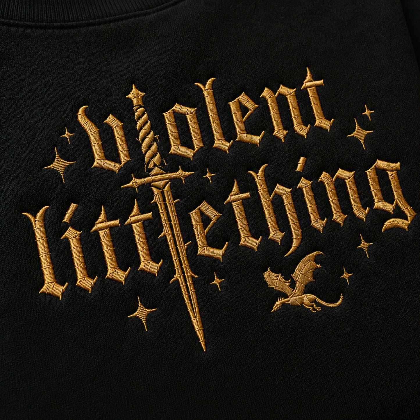 Close-up of a black garment featuring a large gold embroidered gothic-style text design reading 'violent little thing' with a sword, stars, and a small dragon motif.