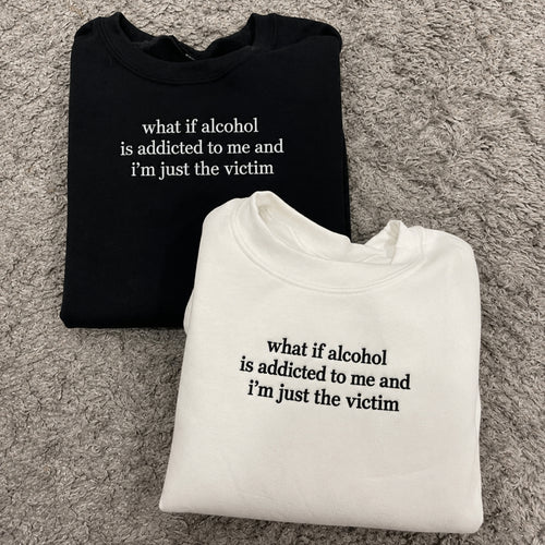 Two folded crewneck sweatshirts on a gray shag rug: one black with white text reading 'what if alcohol is addicted to me and i'm just the victim' and one white with black text reading the same phrase.