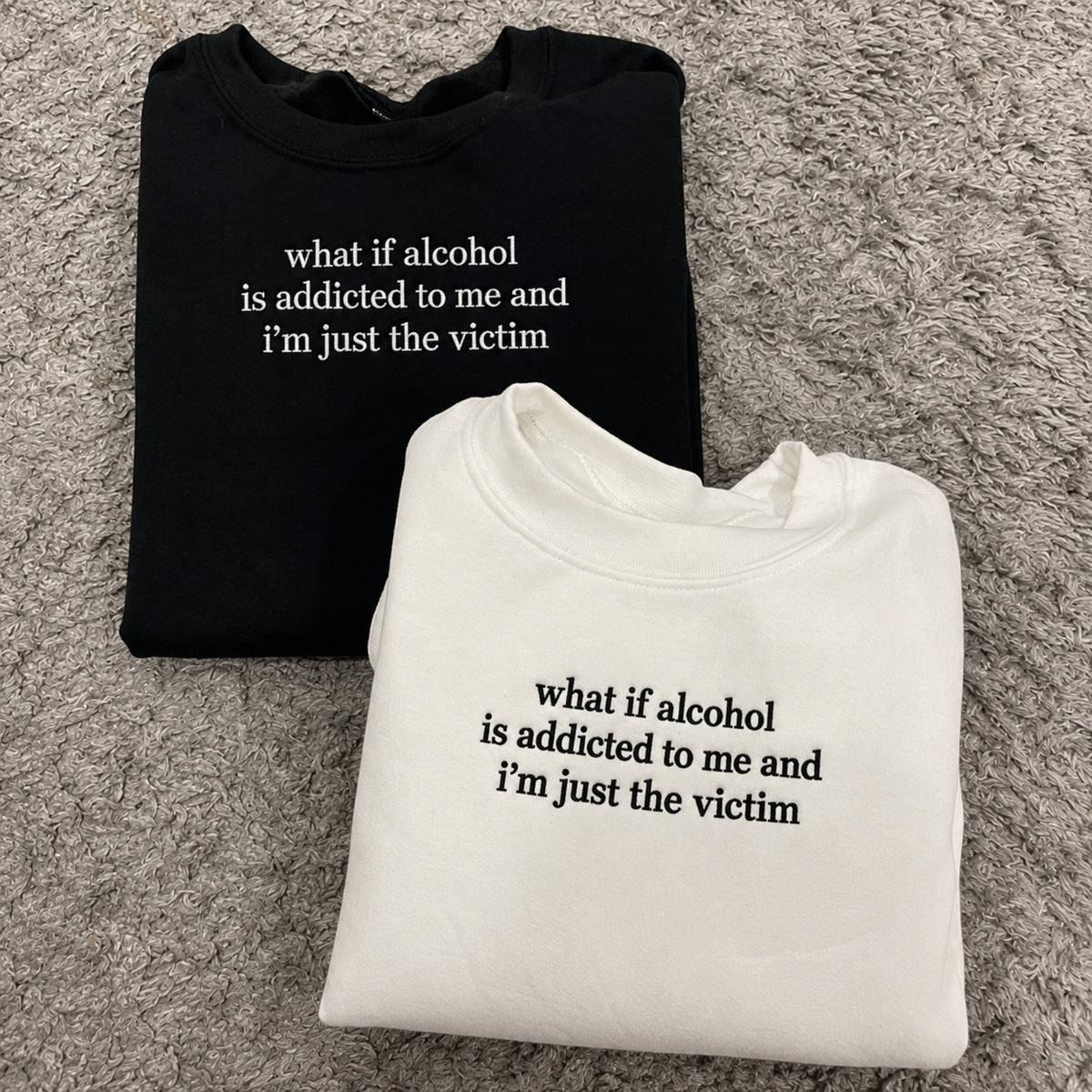 Two folded crewneck sweatshirts on a gray shag rug: one black with white text reading 'what if alcohol is addicted to me and i'm just the victim' and one white with black text reading the same phrase.