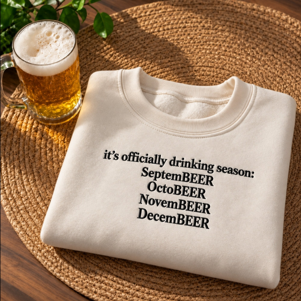 A folded cream sweatshirt with black text reading 'it's officially drinking season: SeptemBEER OctoBEER NovemBEER DecemBEER' sits on a round woven placemat next to a glass of beer.