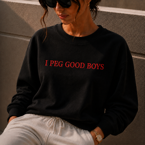 A woman wearing a black crewneck sweatshirt with the phrase 'I PEG GOOD BOYS' printed in red across the chest, paired with white sweatpants, sitting against a textured gray wall.