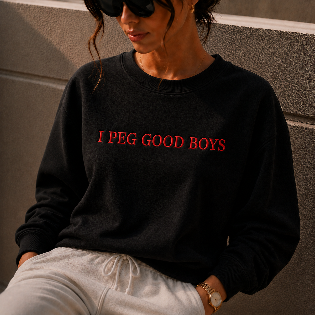 A woman wearing a black crewneck sweatshirt with the phrase 'I PEG GOOD BOYS' printed in red across the chest, paired with white sweatpants, sitting against a textured gray wall.