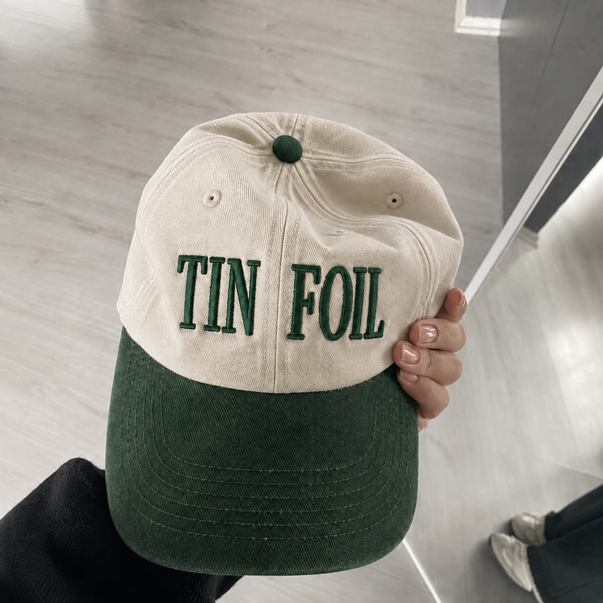 A person holds a two-tone baseball cap with a cream-colored crown and a dark green brim. The front of the crown features the words 'TIN FOIL' embroidered in dark green.