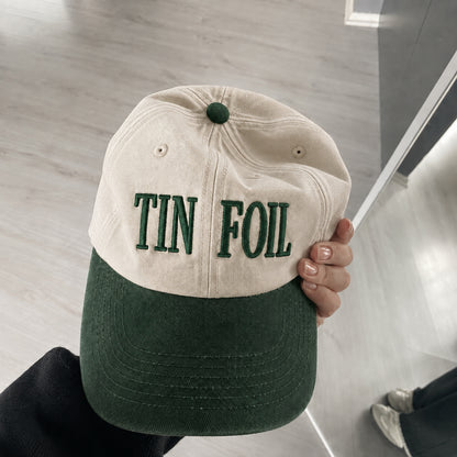 A person holds a two-tone baseball cap with a cream-colored crown and a dark green brim. The front of the crown features the words 'TIN FOIL' embroidered in dark green.