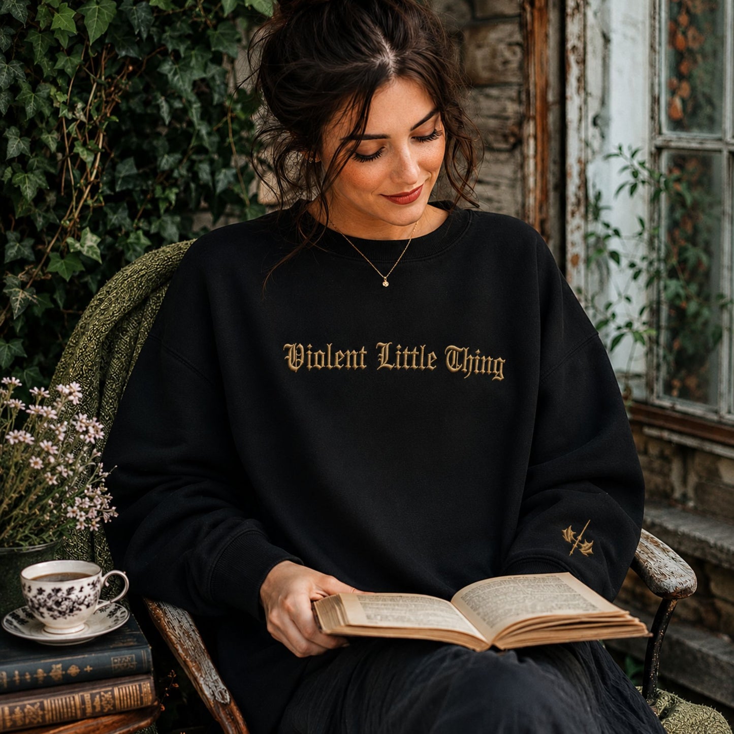 A woman sits in a rustic wooden chair reading an open book, wearing a black crewneck sweatshirt with 'Violent Little Thing' printed in gold gothic lettering across the chest.