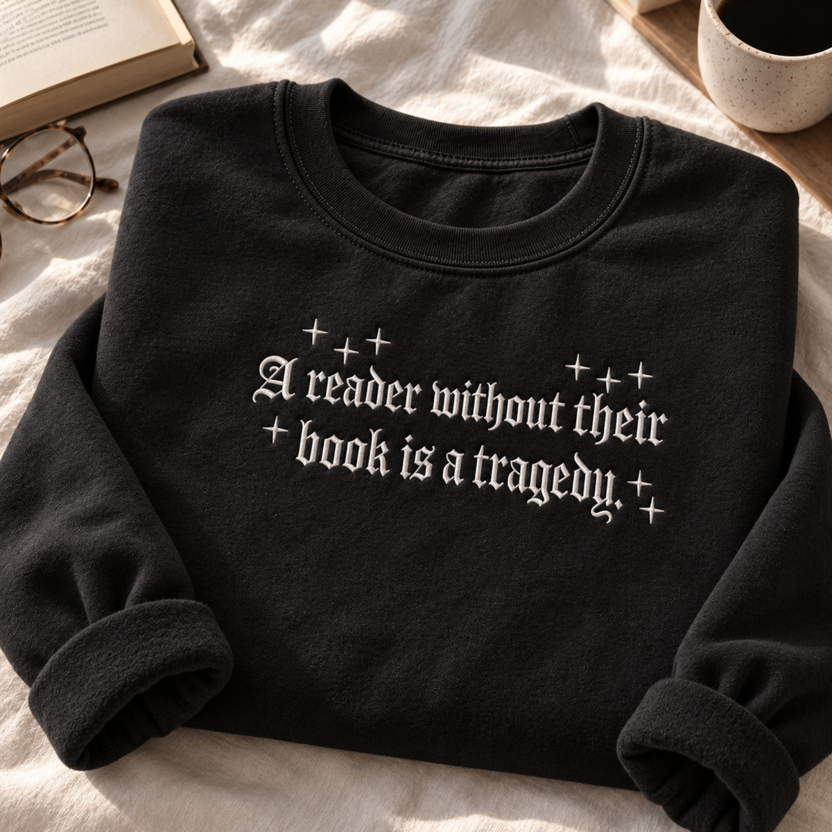 A black crewneck sweatshirt with rolled cuffs, featuring the text 'A reader without their book is a tragedy.' in a white gothic font with decorative plus-sign accents.