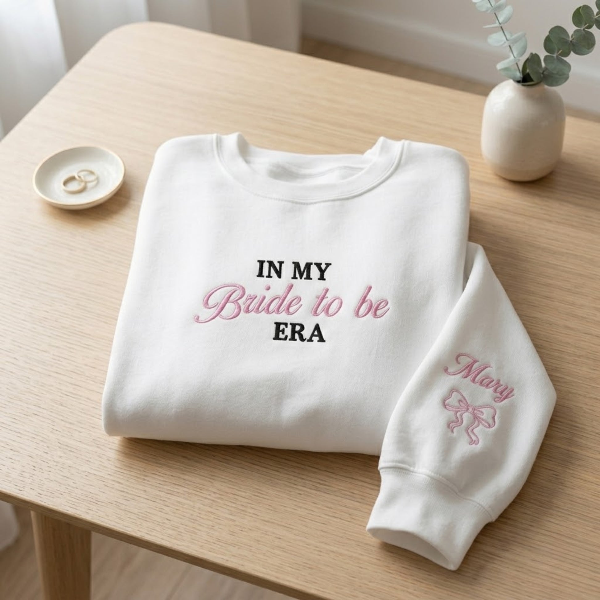 A folded white sweatshirt on a light wood table with embroidered text reading 'IN MY Bride to be ERA' in black and pink, plus a matching pink 'Mary' bow design on the sleeve. Small decorative props, including rings and a small vase, sit nearby.