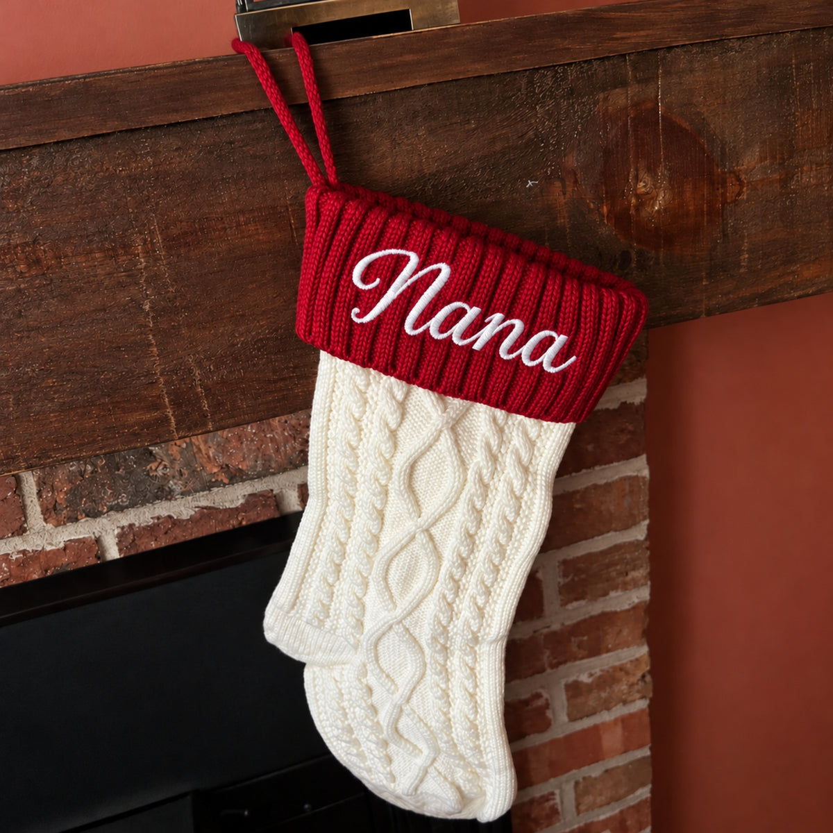A personalized Christmas stocking with a red knit cuff and white cable-knit body, hanging from a dark wooden mantel. The cuff is embroidered with the name 'Nana' in white script.