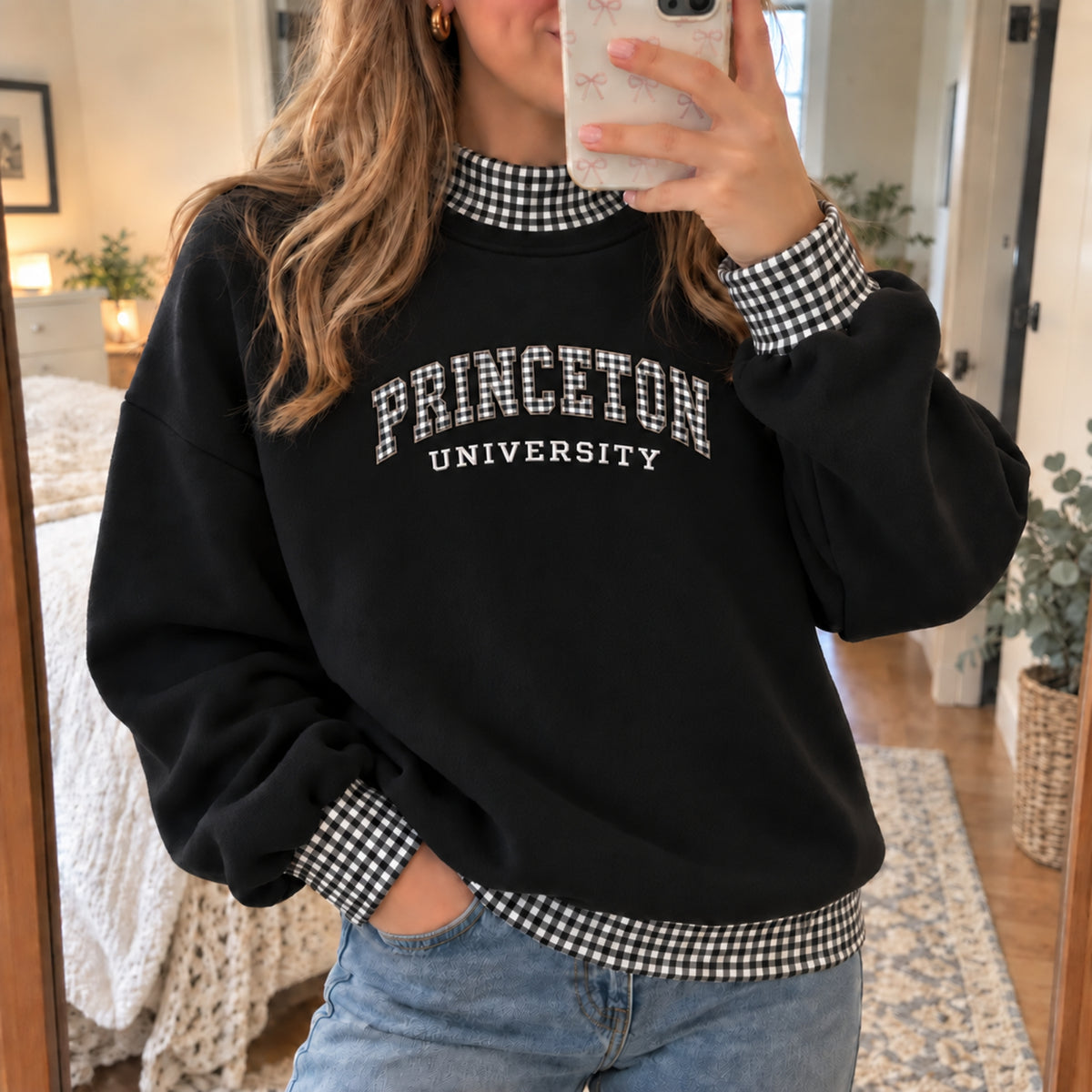 A woman takes a mirror selfie wearing a black crewneck sweatshirt with 'PRINCETON UNIVERSITY' printed in white across the chest. The sweatshirt features a black and white gingham check pattern on the mock neck, cuffs, and hem, and is styled with light blue jeans.