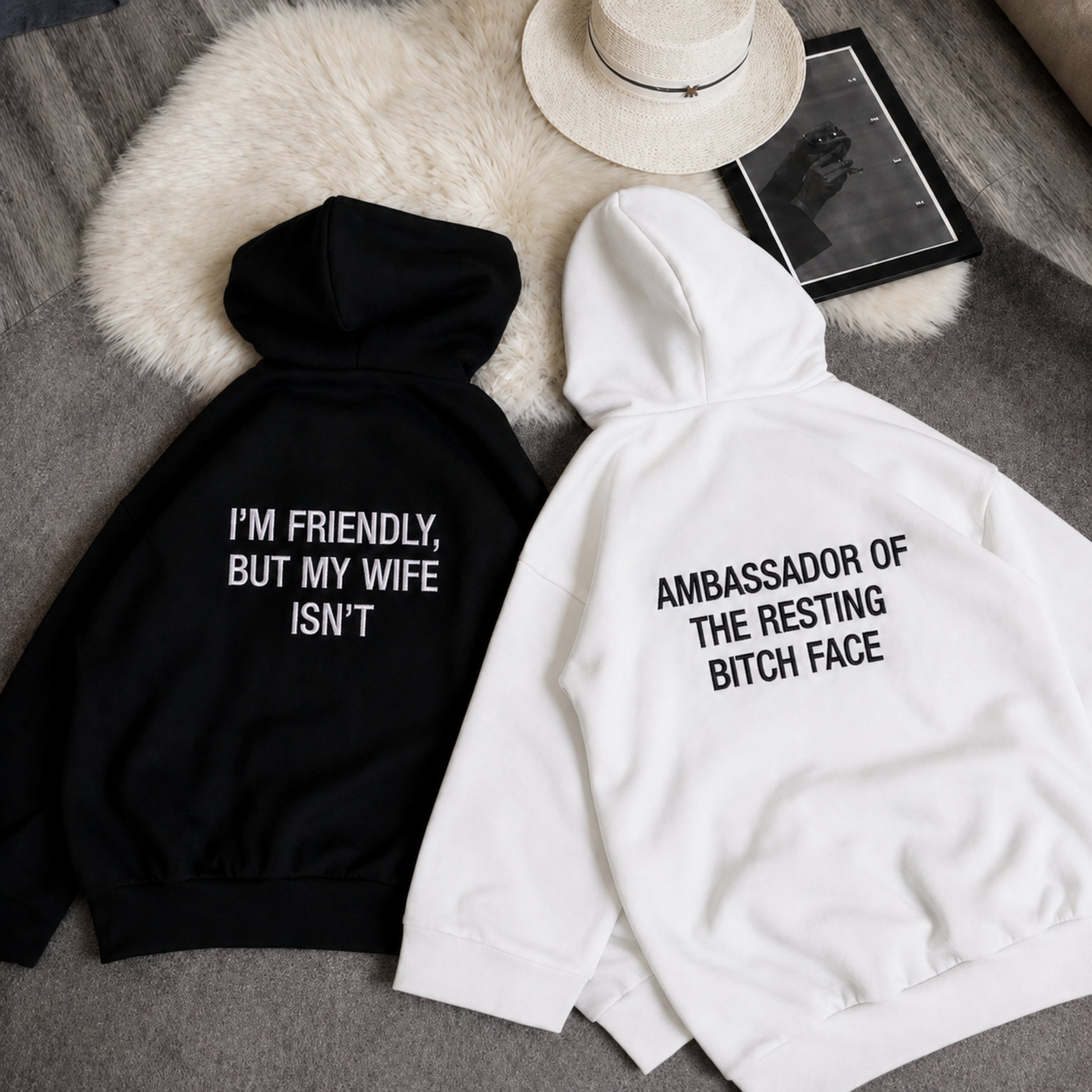 Two hoodies laid flat on a gray fabric surface with a white faux-fur pad and a hat nearby. One hoodie is black with white text reading 'I'M FRIENDLY, BUT MY WIFE ISN'T,' and the other is white with black text reading 'AMBASSADOR OF THE RESTING BITCH FACE.