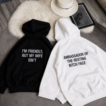 Two hoodies laid flat on a gray fabric surface with a white faux-fur pad and a hat nearby. One hoodie is black with white text reading 'I'M FRIENDLY, BUT MY WIFE ISN'T,' and the other is white with black text reading 'AMBASSADOR OF THE RESTING BITCH FACE.