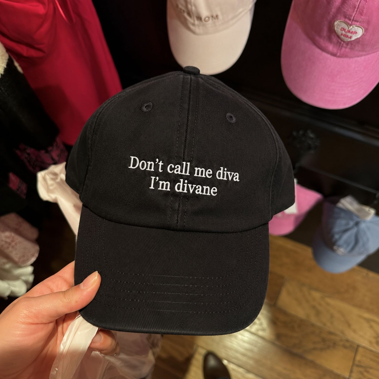 A hand holds a black baseball cap with white embroidered text reading 'Don't call me diva I'm divane'. The background shows a retail display with other colorful caps hanging on a shelf.