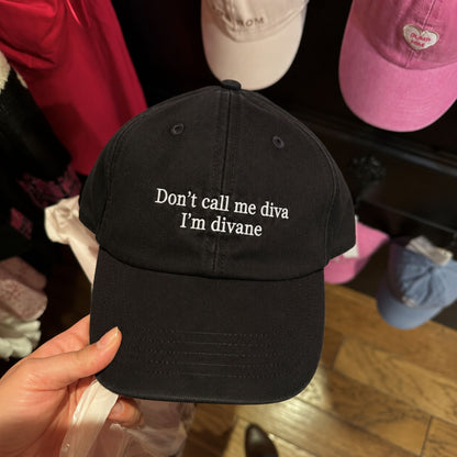 A hand holds a black baseball cap with white embroidered text reading 'Don't call me diva I'm divane'. The background shows a retail display with other colorful caps hanging on a shelf.