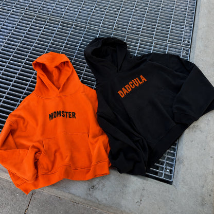 Two hoodies laid flat on a metal grate over concrete: an orange hoodie with black 'MOMSTER' text and a black hoodie with orange 'DADCUVA' text.