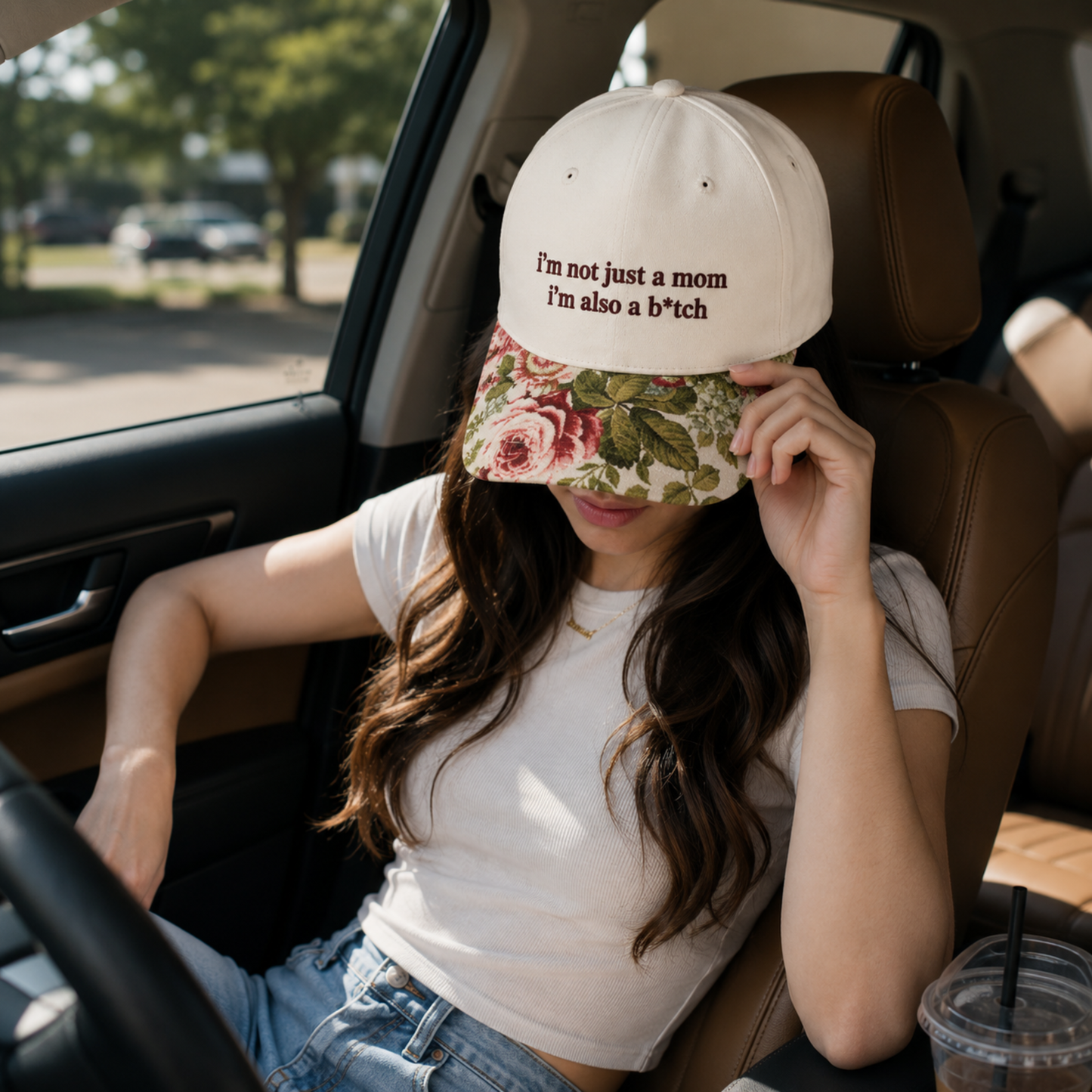 A woman sitting in a car wearing a cream baseball cap with brown text that reads 'I'm not just a mom I'm also a b*tch' and a floral visor. She is wearing a white t-shirt and blue jeans, with a plastic cup on the passenger side.