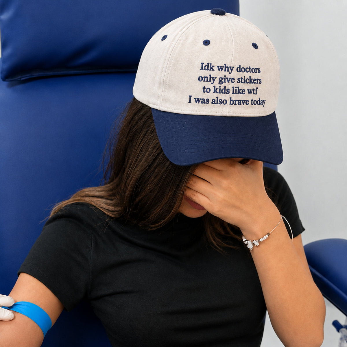 A woman sitting in a blue chair wearing a navy and off-white baseball cap with the text 'Idk why doctors only give stickers to kids like wtf I was also brave today' printed in navy on the front. She is wearing a black shirt and has a blue wristband on her arm.