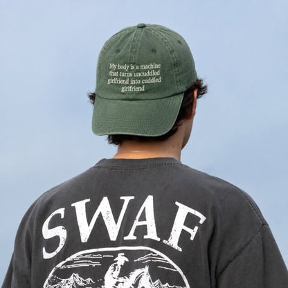 A man viewed from behind wearing an olive green baseball cap with white embroidered text and a faded black t-shirt with a large white graphic on the back. The background is a solid light blue sky.
