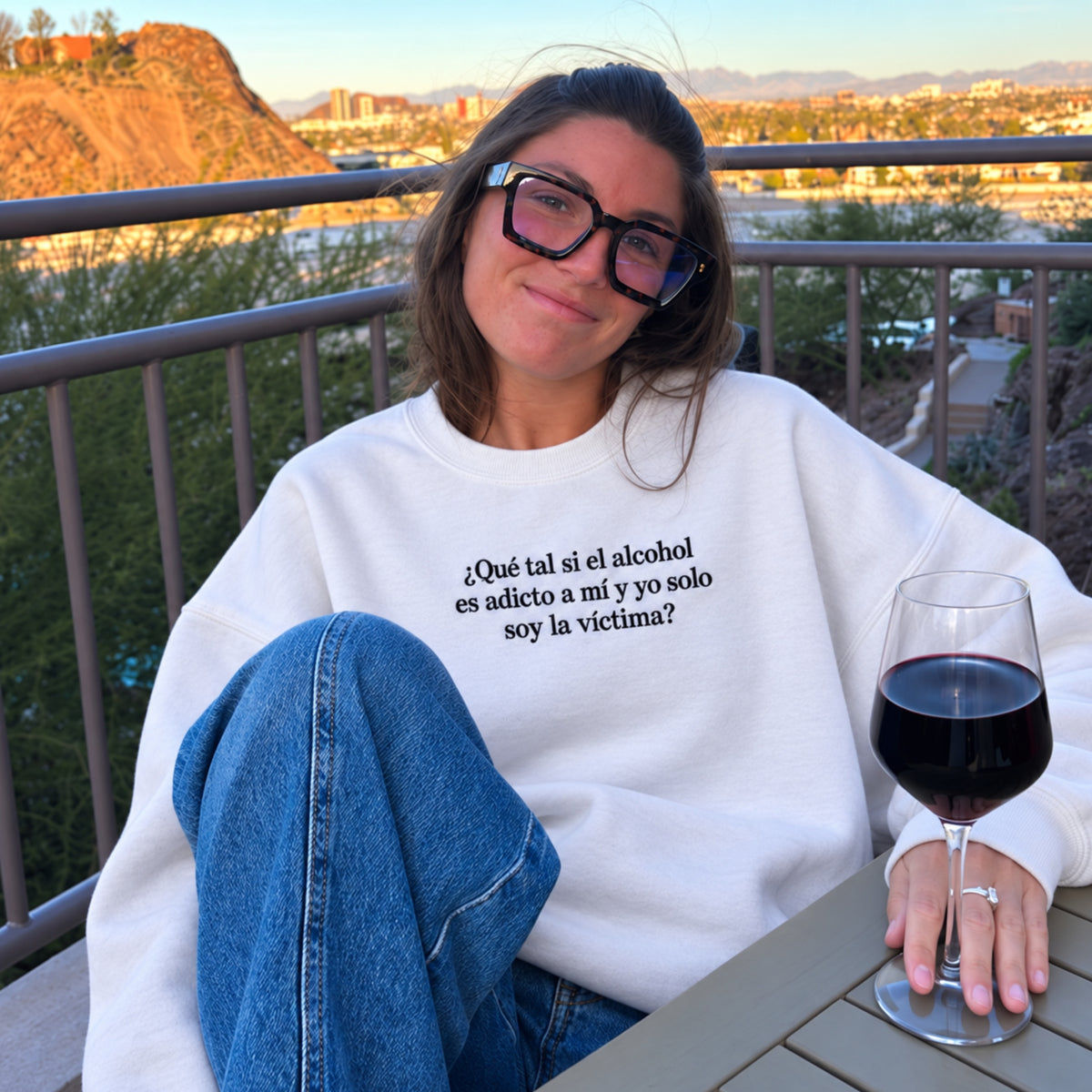 Woman wearing a white embroidered sweatshirt with the Spanish quote “¿Qué tal si el alcohol es adicto a mí y yo solo soy la víctima?” sits outdoors on a balcony beside a glass of red wine.