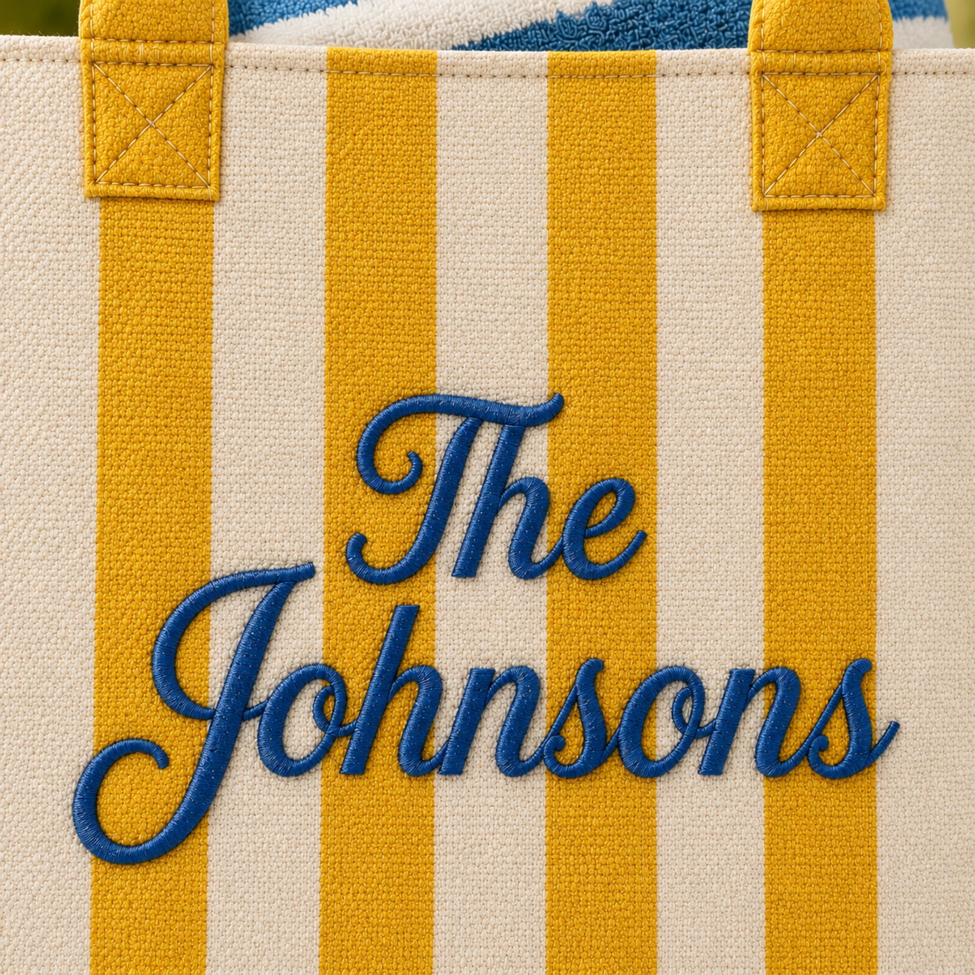 Close-up of a canvas tote bag with vertical yellow and cream stripes, yellow handles, and blue embroidered text reading 'The Johnsons'.