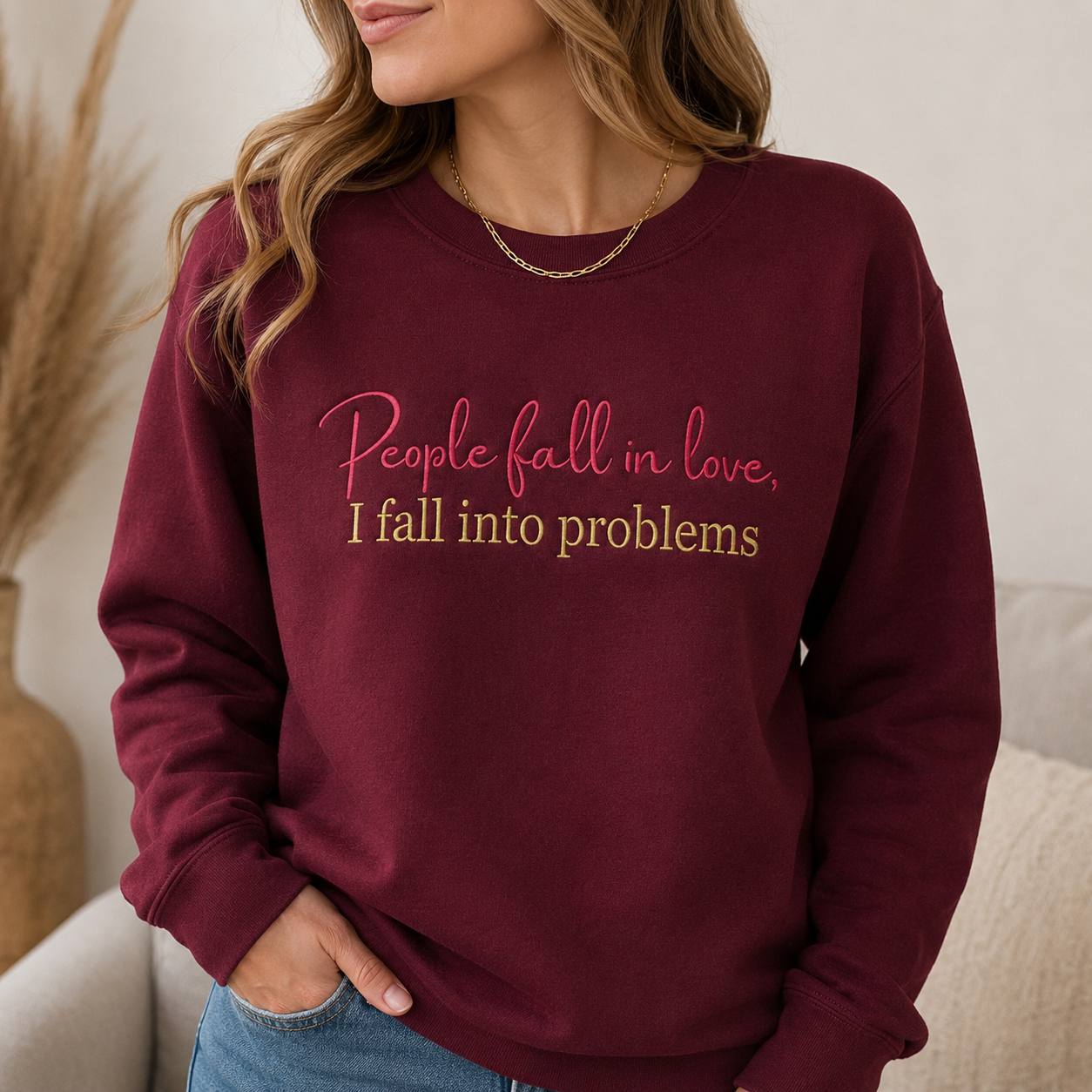 Woman wearing a maroon crewneck sweatshirt embroidered with pink cursive text reading "People fall in love," above gold block lettering reading "I fall into problems."