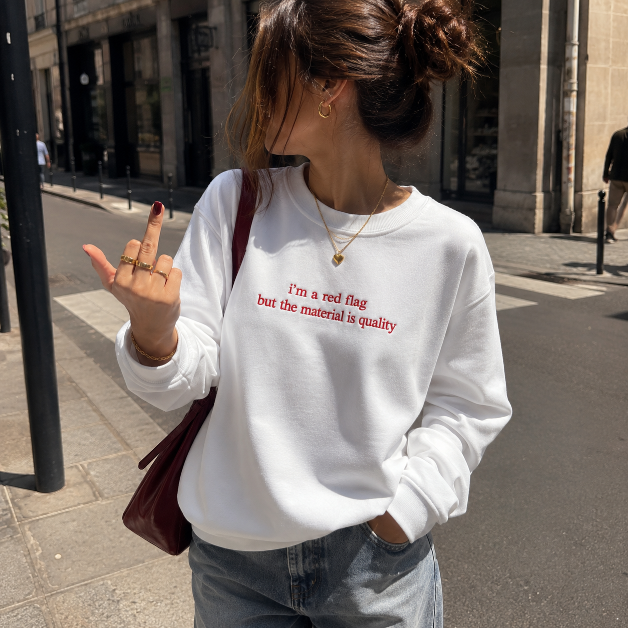 Woman standing on a city sidewalk wearing a white crewneck sweatshirt embroidered with red text reading "i'm a red flag but the material is quality".