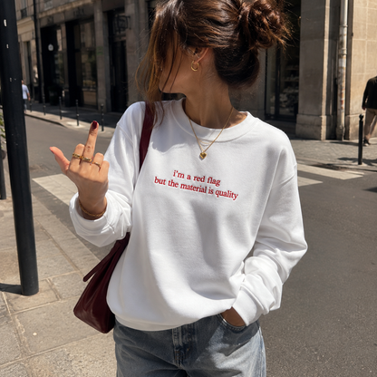 Woman standing on a city sidewalk wearing a white crewneck sweatshirt embroidered with red text reading "i'm a red flag but the material is quality".