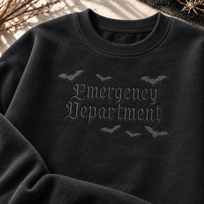 A close-up of a black crewneck sweatshirt featuring a dark gray gothic-style embroidered design that reads 'Emergency Department' with bats flying above and below. The sweatshirt is styled with autumn-themed props like dried leaves and a spider on a textured background.