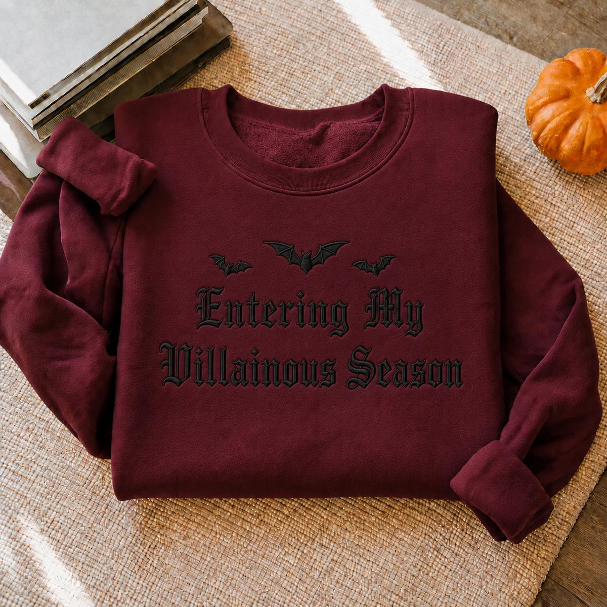 A folded burgundy sweatshirt with black gothic text reading 'Entering My Villainous Season' and three bat silhouettes, styled on a woven beige surface with a small orange pumpkin and books nearby.